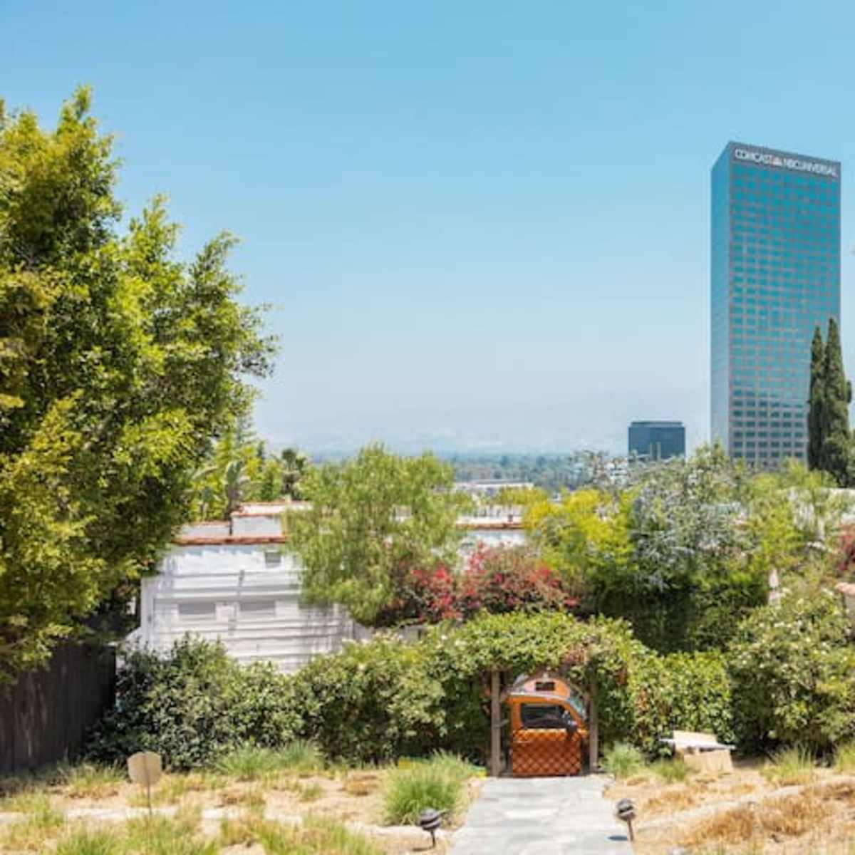 A suburban landscape with a stone pathway leading to a wooden gazebo, surrounded by greenery, with tall buildings visible in the background.