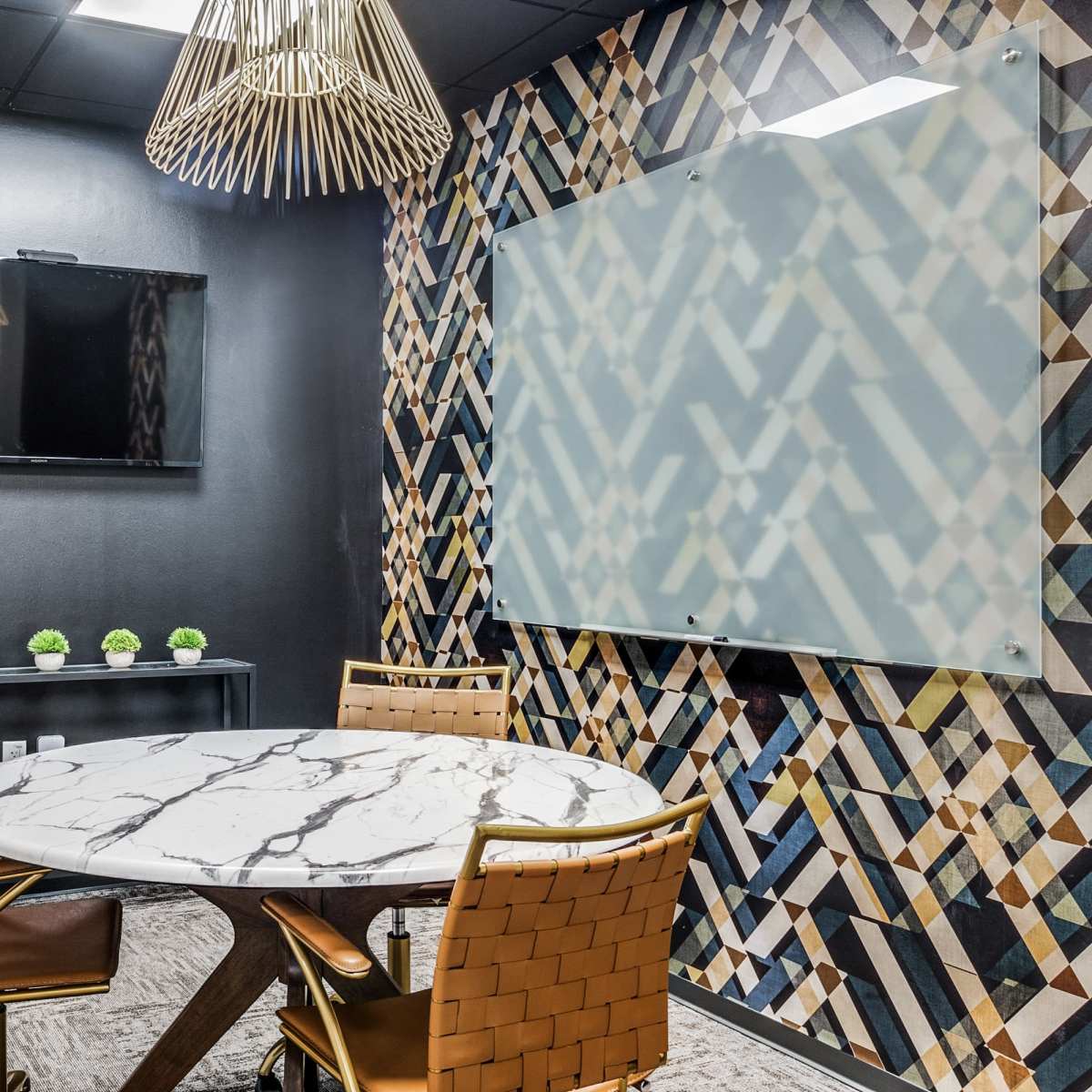 A modern meeting room features a round marble table with gold chairs, a flat-screen TV on the wall, and a decorative patterned backdrop.
