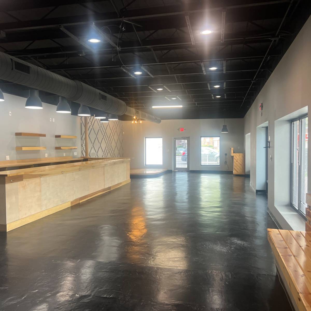 A spacious, empty commercial interior with polished black floors, a long wooden counter, and large windows letting in natural light.