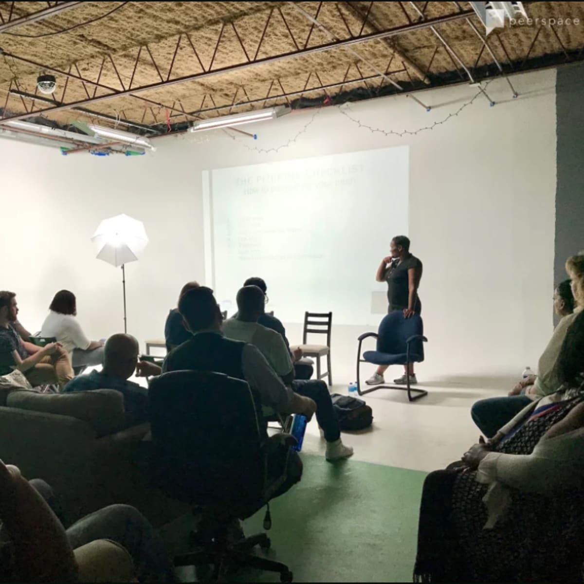 A speaker presents in front of an audience seated on various chairs and couches in a studio with a green screen backdrop.