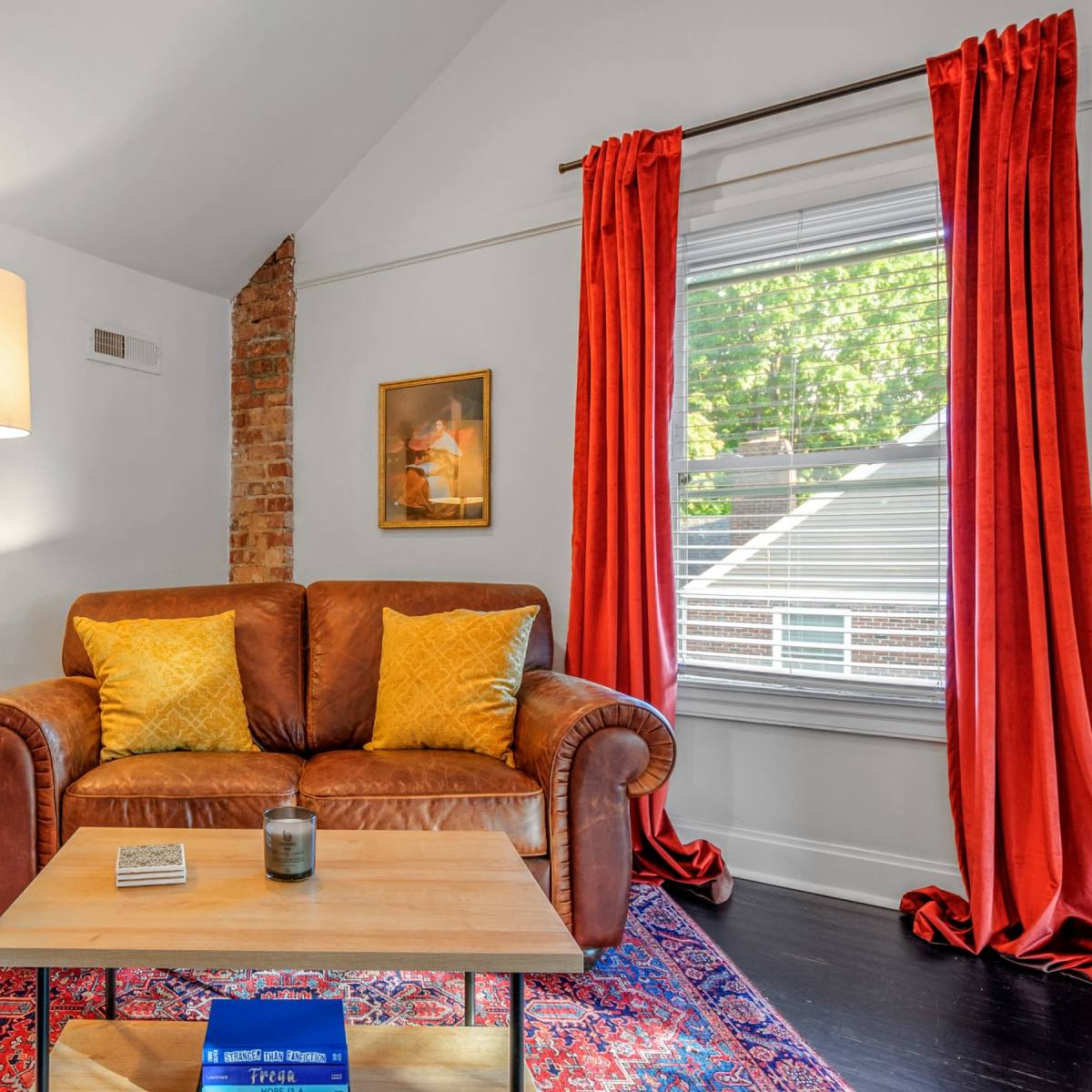 A living room features a brown leather sofa with yellow cushions, a wooden coffee table, a floor lamp, and red curtains framing a window.