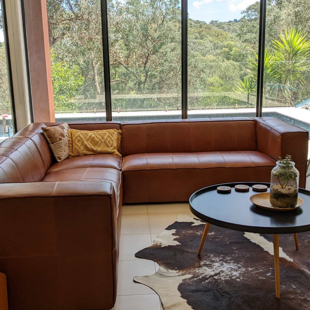 A modern living room features a brown leather sectional sofa, a round coffee table on a cowhide rug, and large windows overlooking a green landscape.