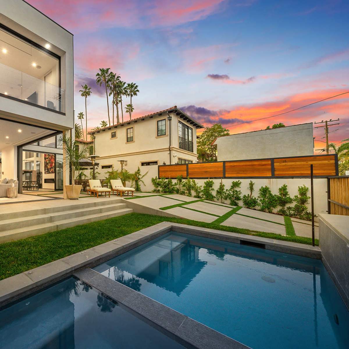 A modern house with large glass windows is surrounded by a neatly landscaped yard and a small swimming pool, set against a colorful sunset.