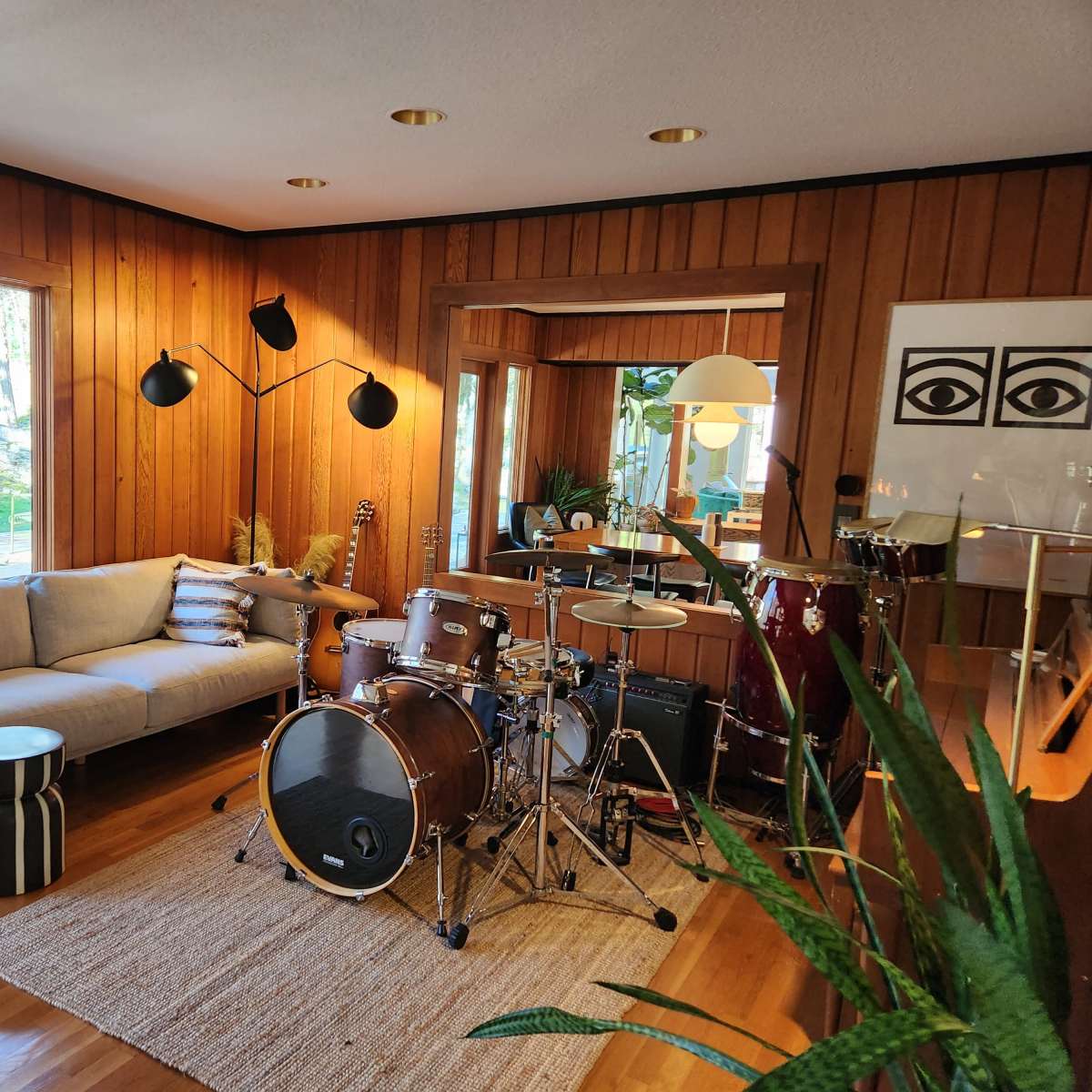 A living room with wooden walls, a couch, a coffee table, a drum set, and a large window that overlooks a garden.