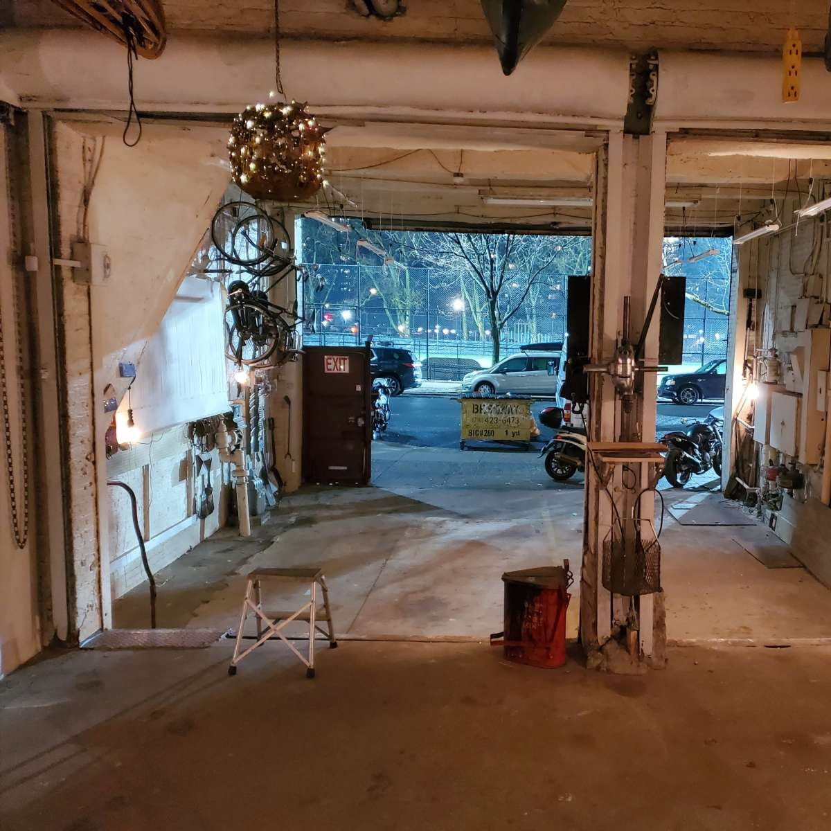 An open garage space with visible tools and equipment on the walls, leading to a view of a street with parked cars in the background.