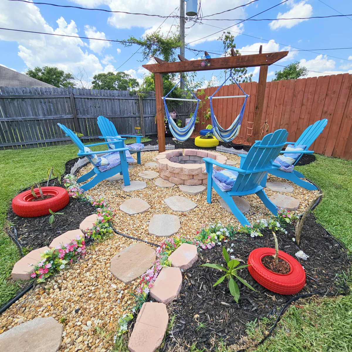 A landscaped backyard area featuring blue Adirondack chairs, a stone fire pit, and hanging hammocks, surrounded by a pathway of stones and flowers.