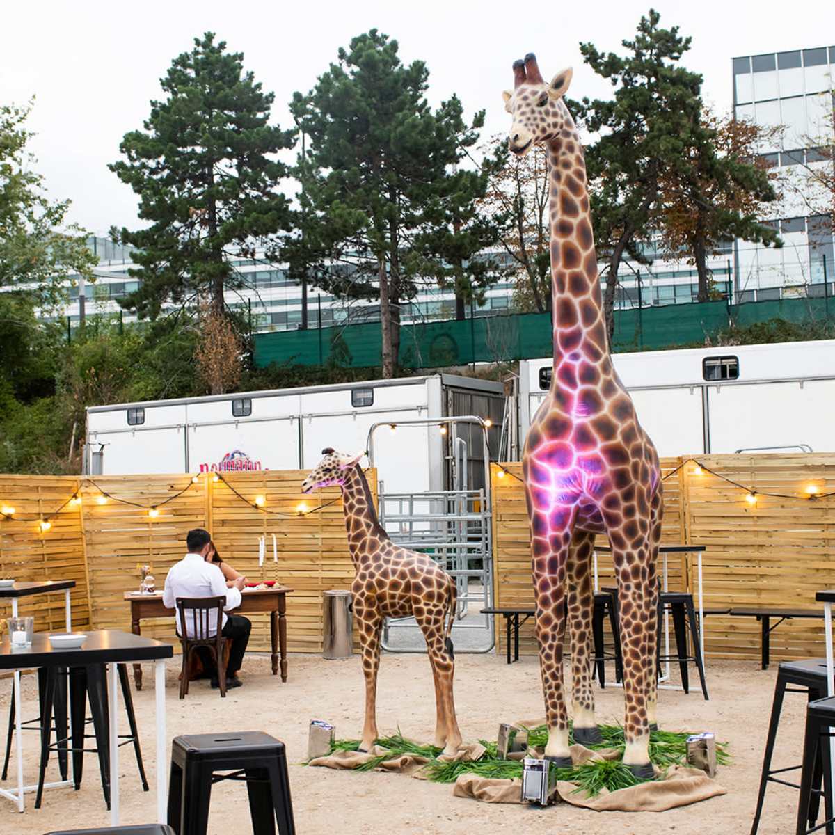 The scene features a fenced outdoor space with two large giraffe statues, surrounded by tables and chairs, and a person sitting nearby.