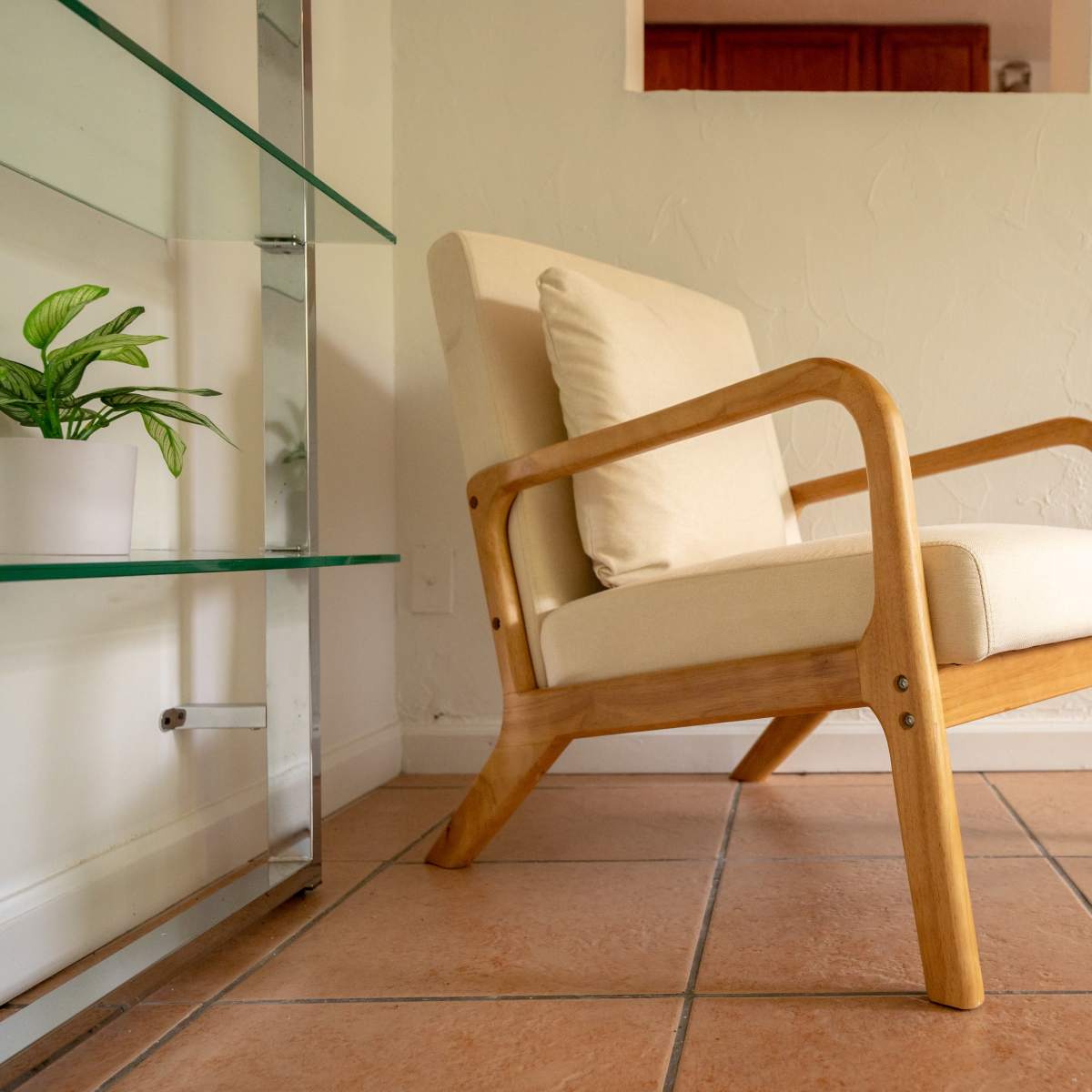 A wooden armchair with cream-colored cushions sits in a room featuring tiled flooring and a small plant on a glass shelf nearby.