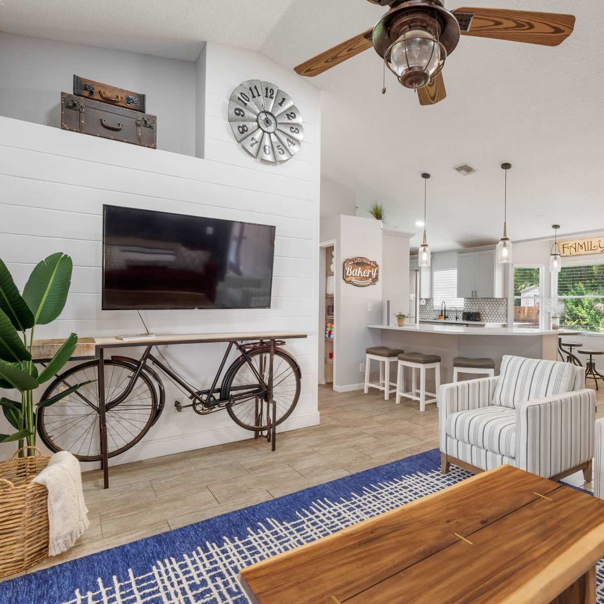 A modern living area featuring a television mounted on a white panel, a decorative bicycle, two striped chairs, a wooden coffee table, and a kitchen area in the background.