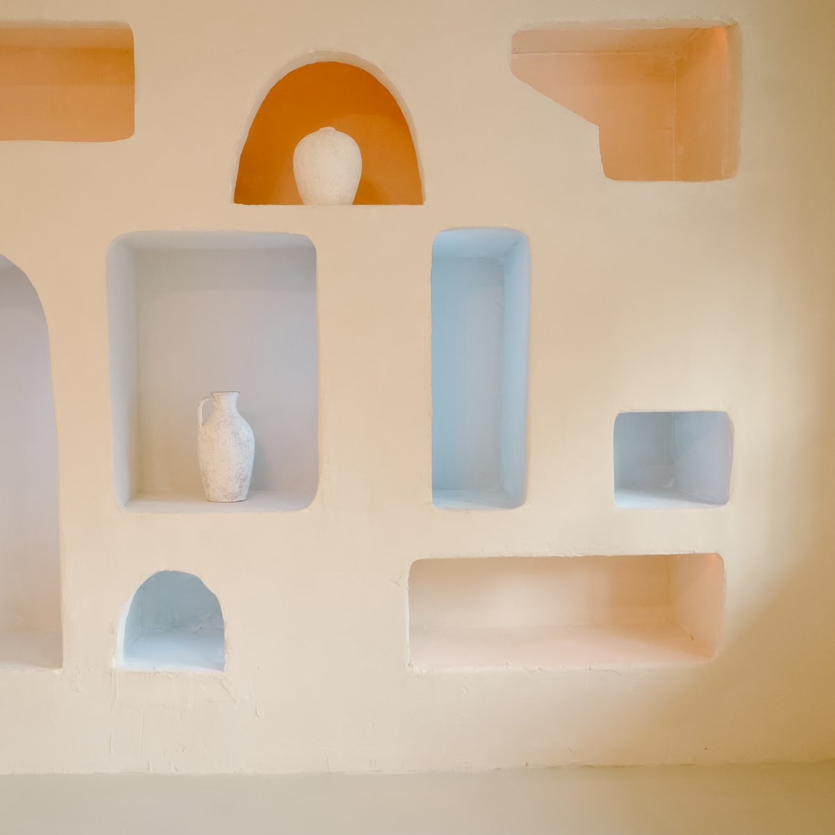 A beige wall with several recessed alcoves in various shapes, displaying a white vase and other decorative elements.
