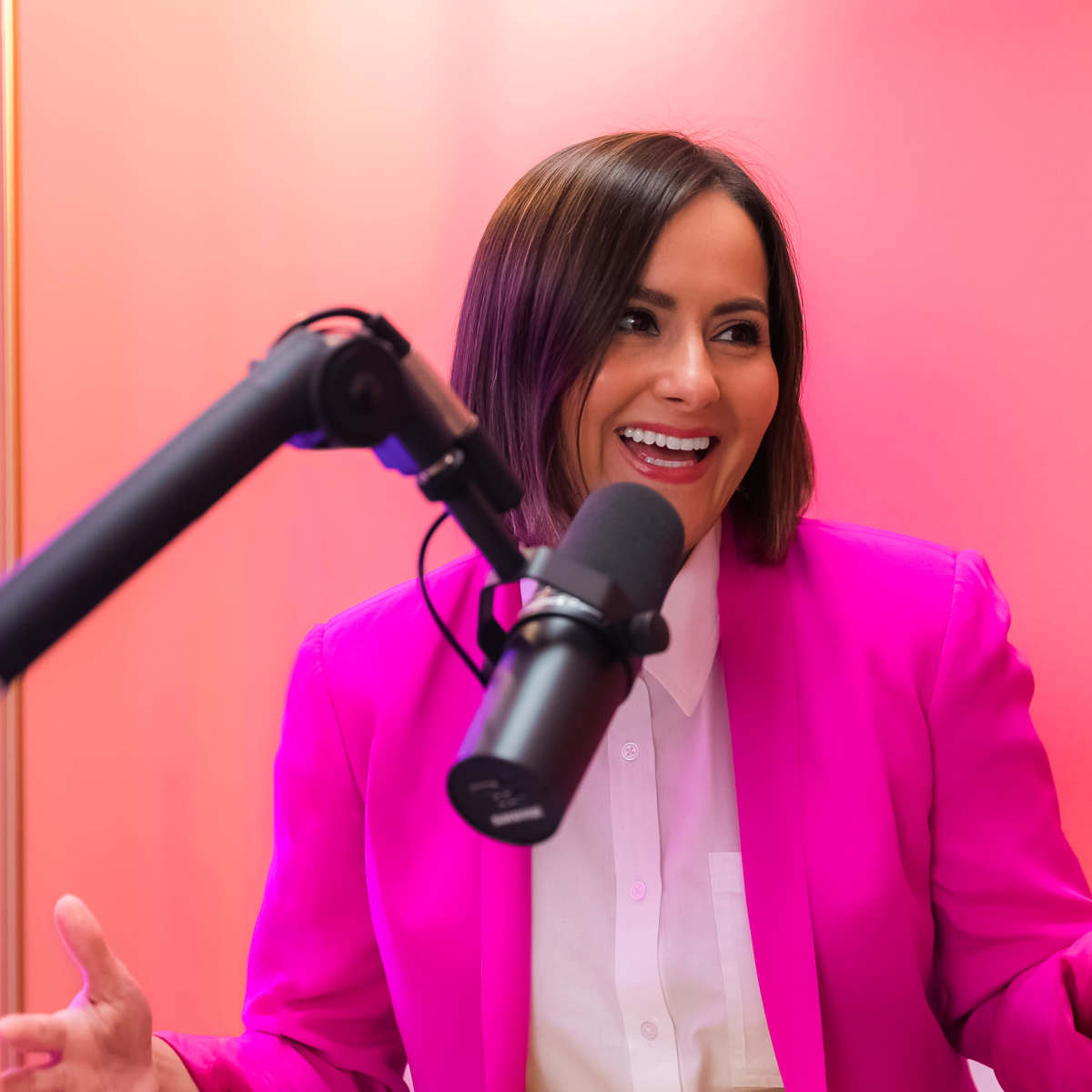 A woman in a bright pink blazer sits in front of a microphone, gesturing as she speaks against a pink background.
