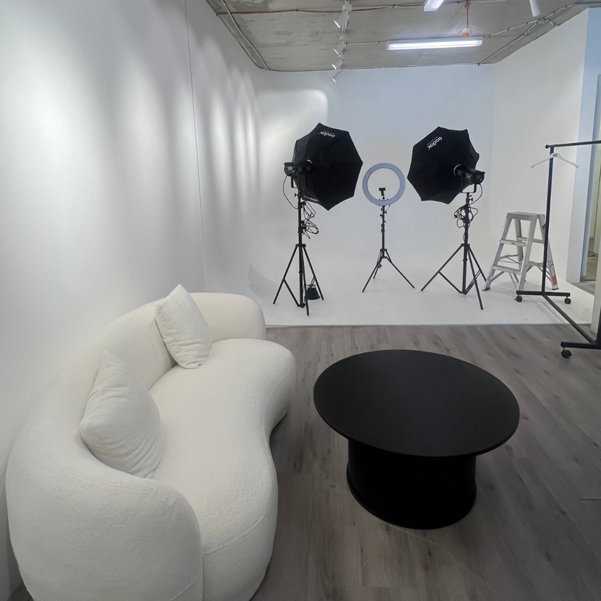 A photography studio featuring a white curved couch and a black circular coffee table, with two softbox lights positioned on tripods against a white backdrop.