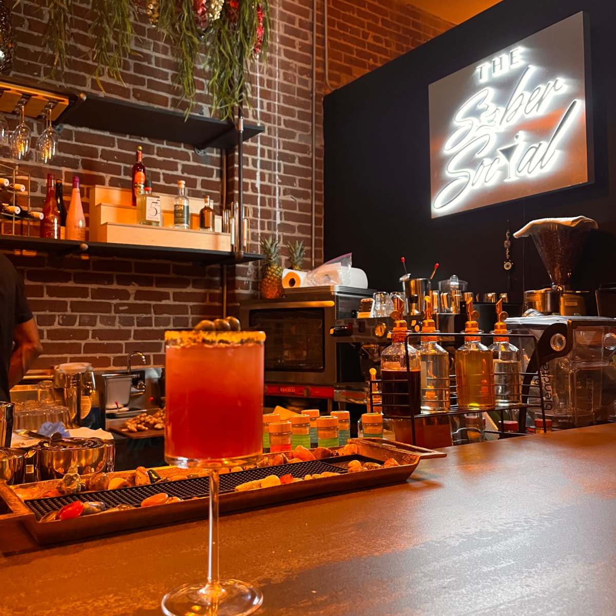 A dimly lit bar scene with a cocktail on the counter in the foreground, and a stylish bar setup featuring shelves of liquor and a neon sign in the background.
