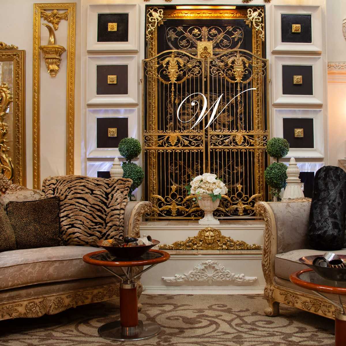 An opulent living room featuring ornate gold decor, two elaborately designed sofas, and a decorative wall with a monogrammed gate.
