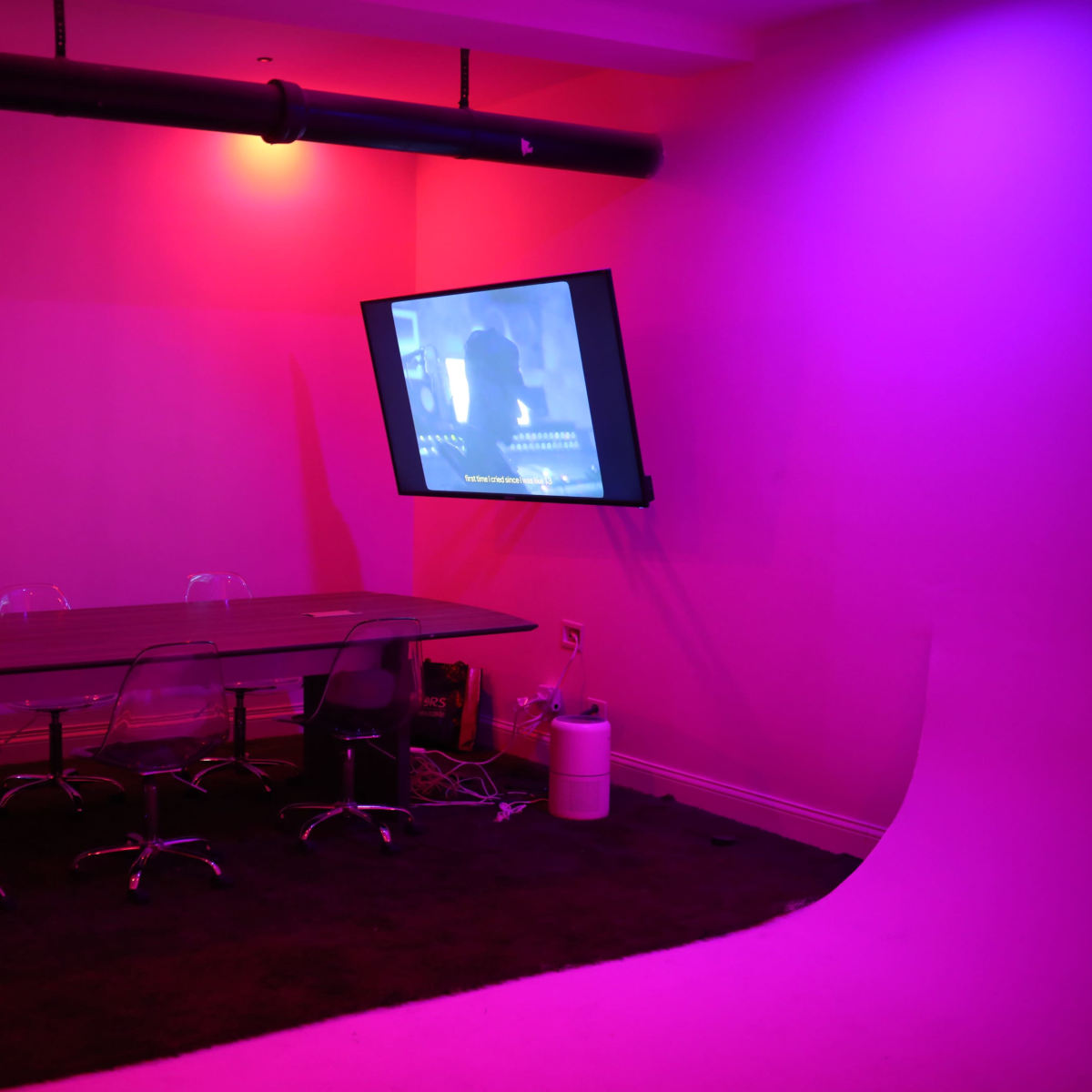A dimly lit room features a large rectangular table surrounded by clear chairs, with a wall-mounted screen displaying a projection.