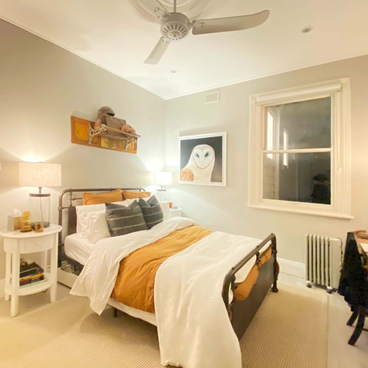 A neatly arranged bedroom features a bed with orange and white bedding, a bedside table, a window with drapes, and artwork of an owl on the wall.