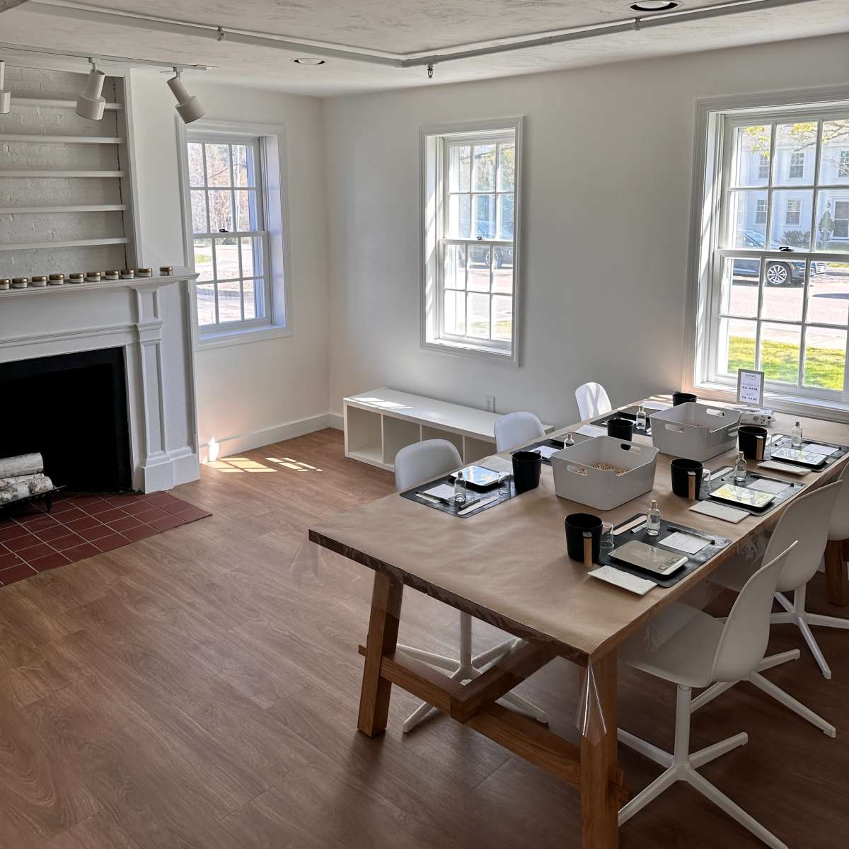 A bright, minimalist dining area with a large wooden table set for a meal, surrounded by white chairs and natural light pouring in through adjacent windows.