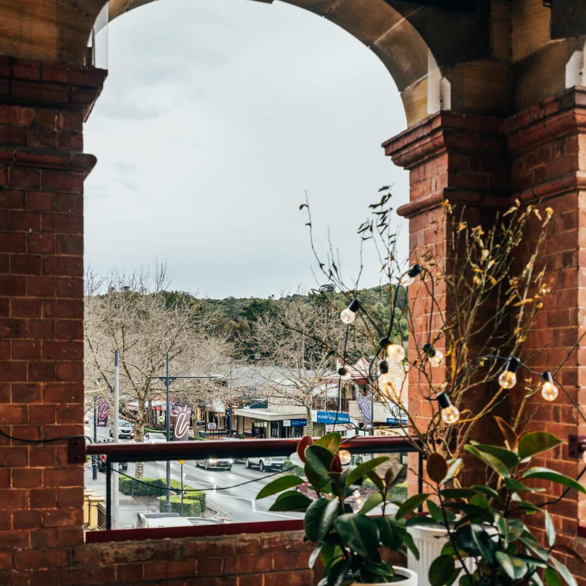 A stone archway frames a view of a street lined with bare trees and shops, with potted plants positioned in the foreground.