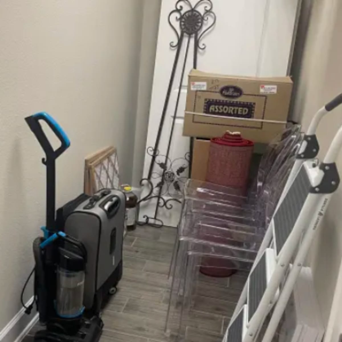 A narrow storage area contains a vacuum cleaner, a ladder, stacked chairs, and several boxes labeled "Assorted."