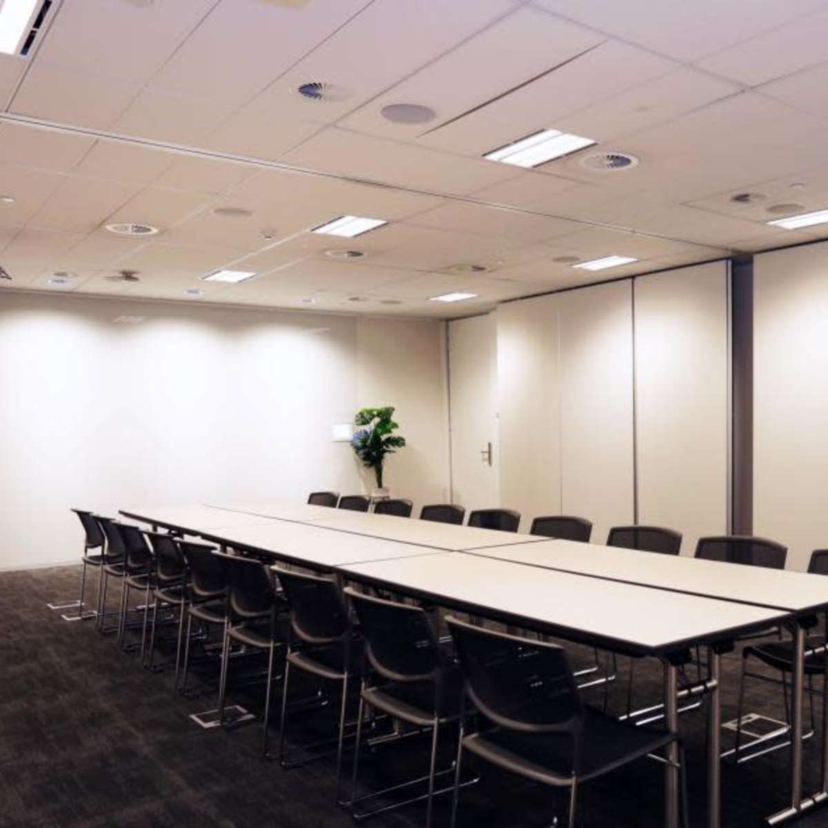 A long conference table with black chairs is set up in a modern meeting room featuring sliding wall panels and potted plants in the corners.