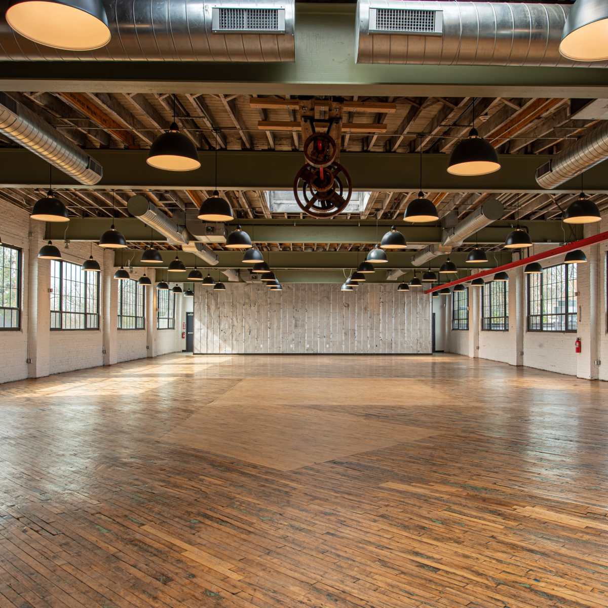 A spacious, well-lit industrial room with large windows and wooden floors, featuring exposed beams and overhead lighting.
