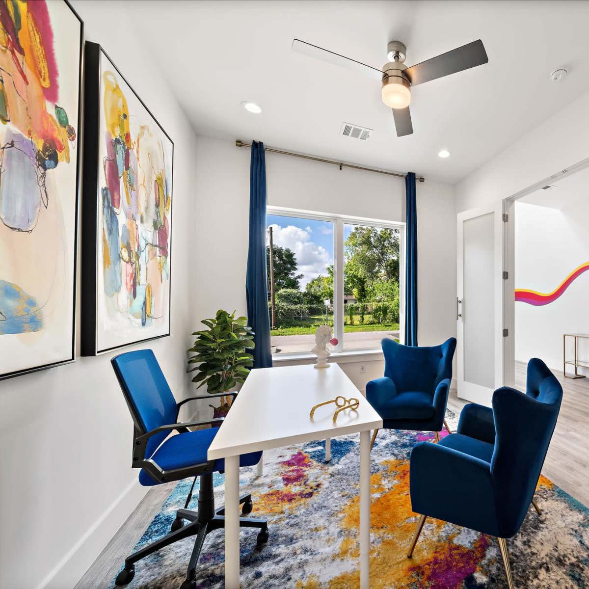 A brightly lit office space features a white table, two blue chairs, colorful artwork on the walls, and a large window with a view of greenery.