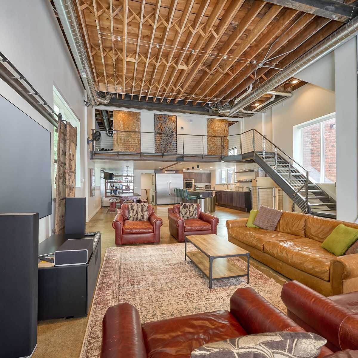 A spacious, modern living area with exposed wooden beams, multiple seating options, and a visible kitchen area in a loft-style design.