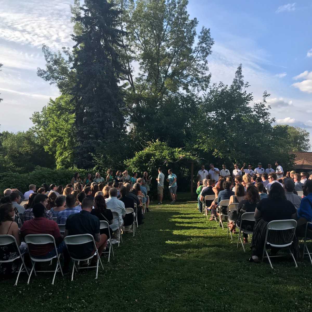 A large group of people is gathered on chairs in a grassy area for a wedding ceremony, with trees and a blue sky in the background.