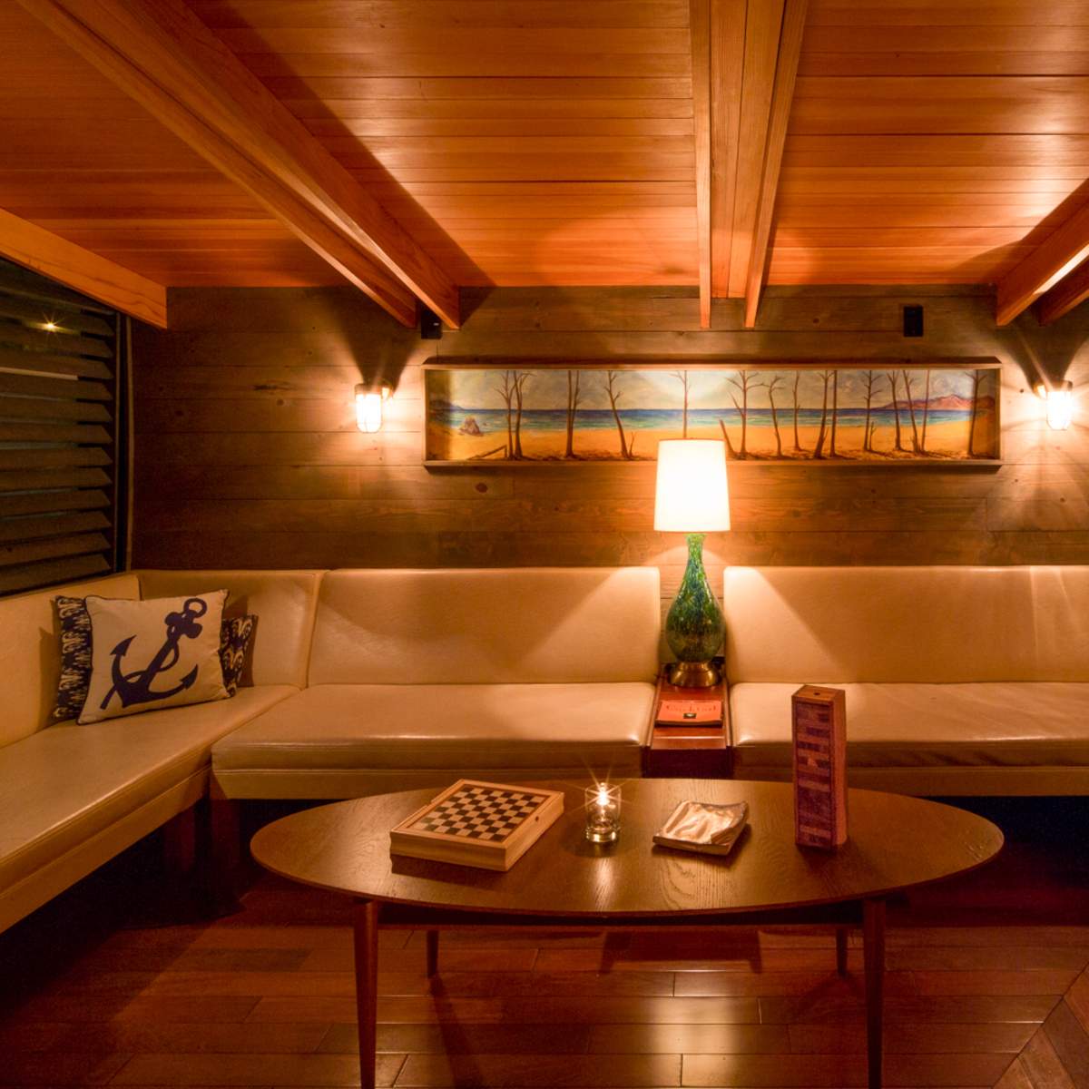 A cozy indoor seating area features a sectional couch with decorative pillows, a wooden table, and a lamp illuminating a wall art piece above.