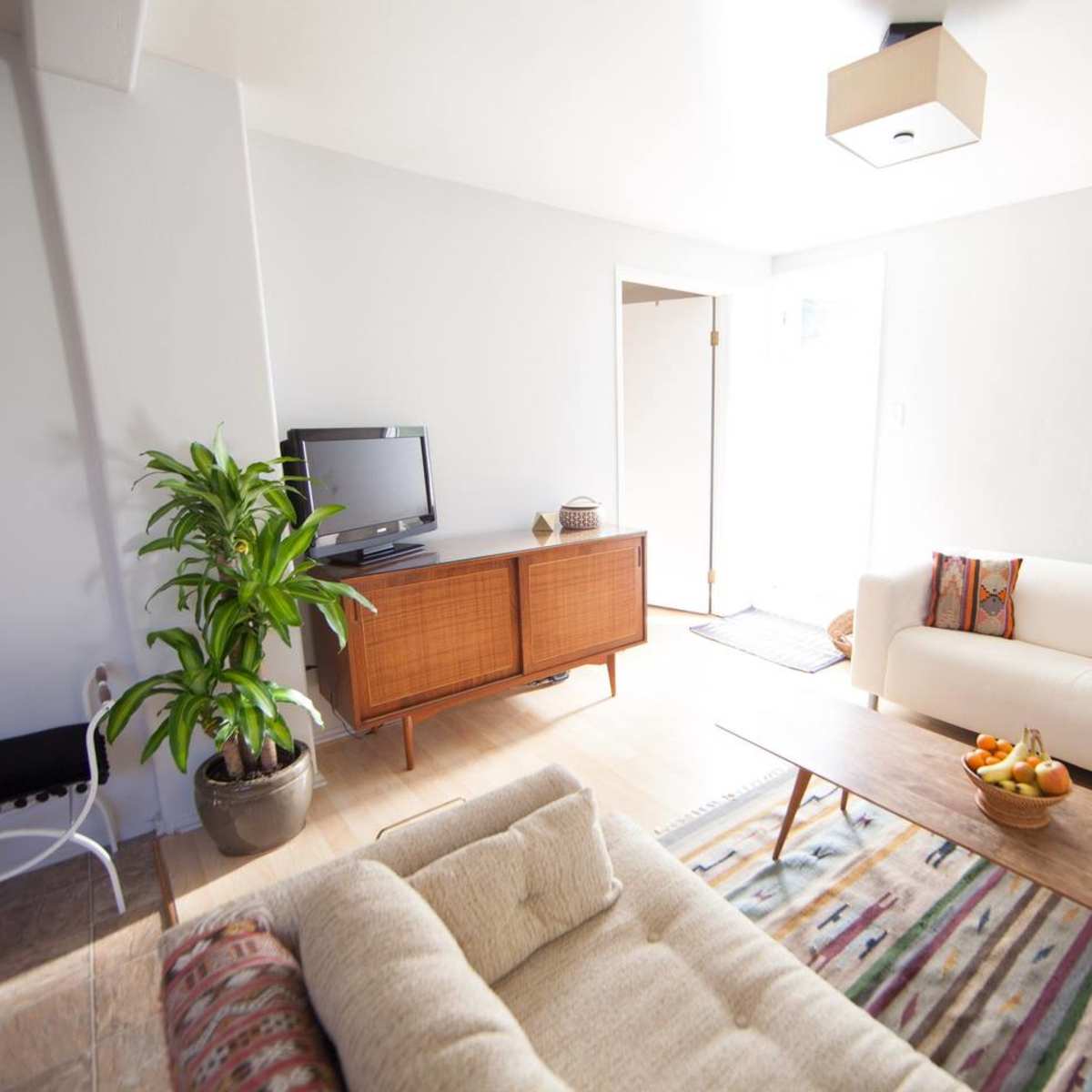 A light-colored living room with a white sofa, a wooden coffee table, a television on a cabinet, and a decorative plant in the corner.