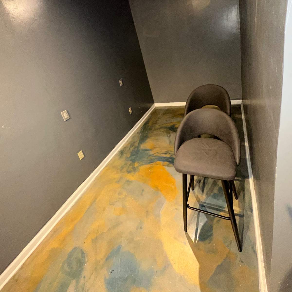 A small, empty room with gray walls and two gray bar stools positioned on a floor with a blue and orange pattern.