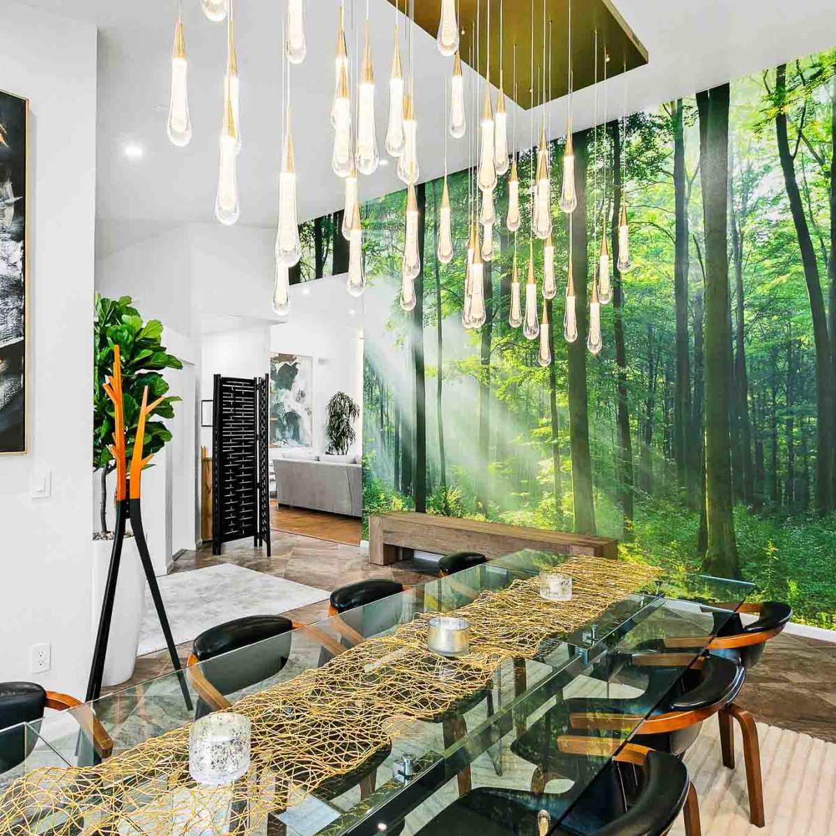 A modern dining area features a long glass table set with decorative elements, surrounded by black chairs, with a forest-themed mural on one wall and unique pendant lighting above.
