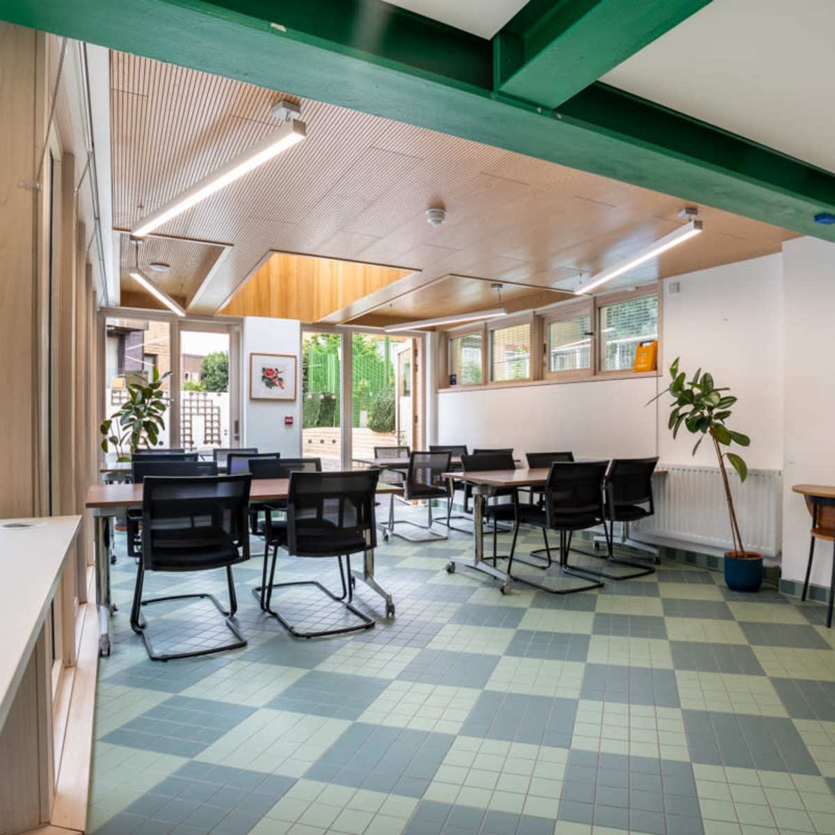 A well-lit multi-purpose room with tables and chairs arranged for meetings, featuring large windows overlooking a garden.