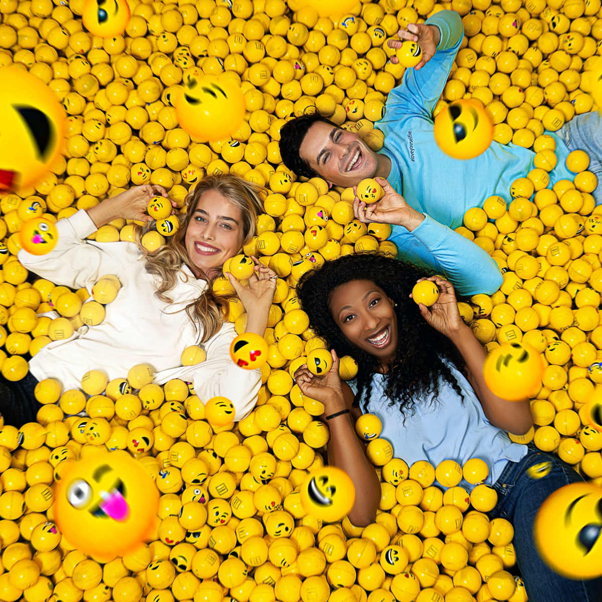 Three people lie in a pool of yellow balls decorated with emoji faces, smiling and posing for the camera.
