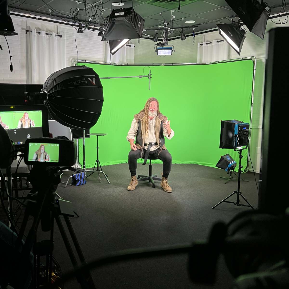 A man with long hair and a beard sits on a stool in front of a green screen while being filmed by multiple cameras in a studio.