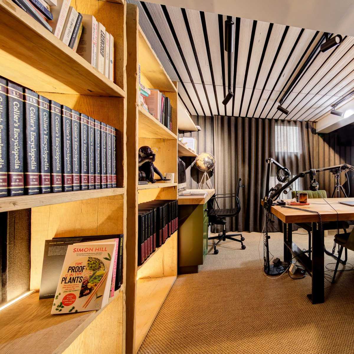 A modern office space featuring a wooden bookshelf filled with books, a large desk with two chairs, and various desk lamps under striped ceiling panels.