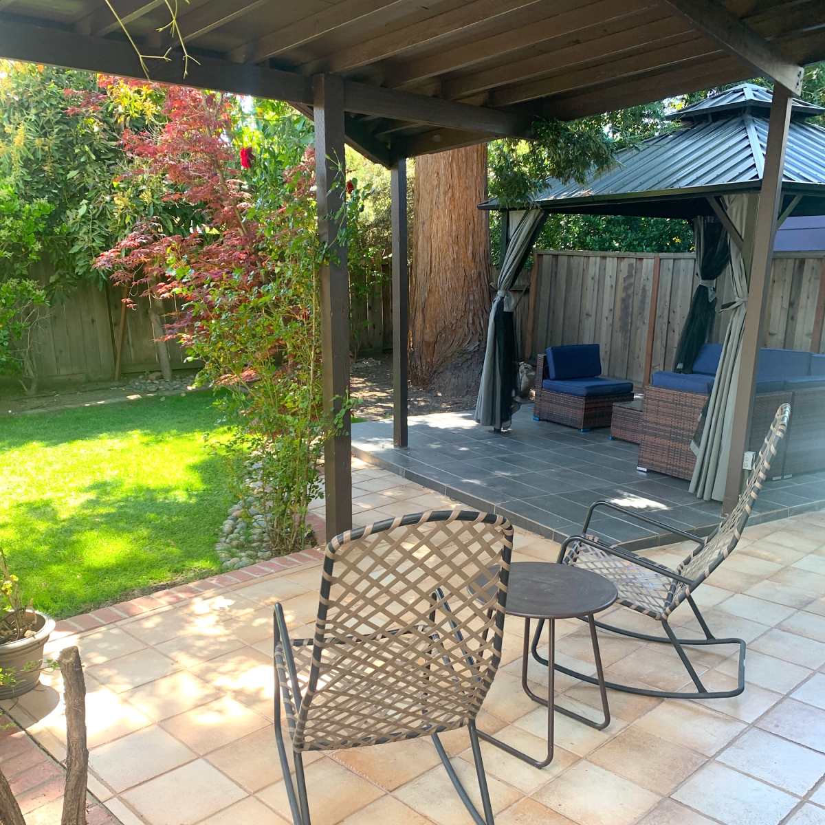 A garden area with two rocking chairs and a small table under a pergola, overlooking a lawn and a seating area with a gazebo in the background.