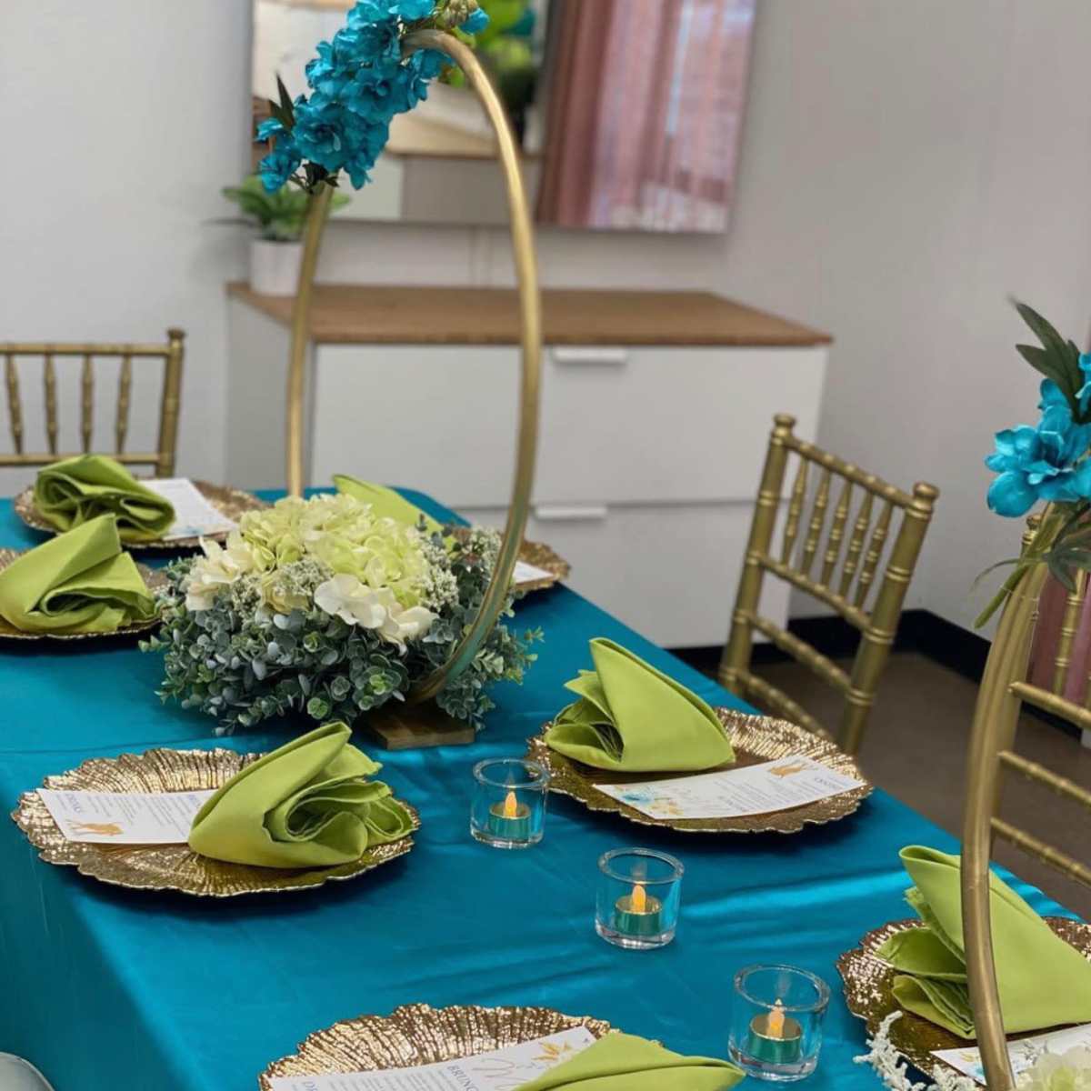 A neatly arranged dining table features gold chargers, green napkins, and a floral centerpiece, with gold chiavari chairs surrounding the setup.