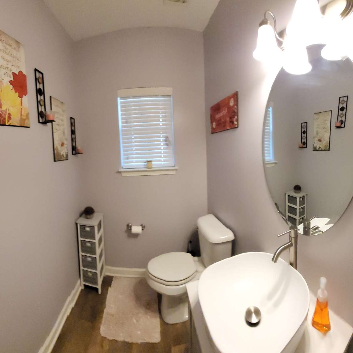 A small bathroom with light purple walls, a round mirror above a sink, a toilet, a small storage cabinet, and decorative wall art.