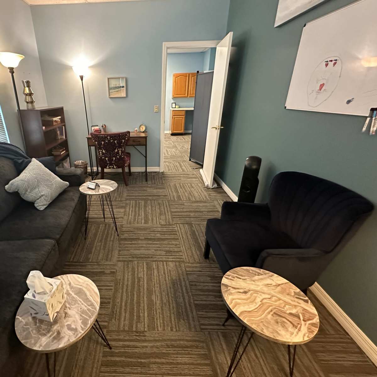 A cozy room with a dark sofa, two small marble tables, a chair, and a door leading to a kitchen area.