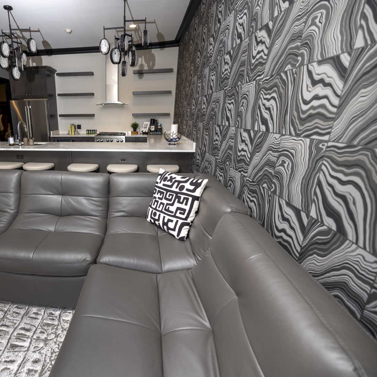 A modern living room features a gray sectional sofa, a patterned area rug, and a kitchen counter with sleek cabinetry and appliances in the background.