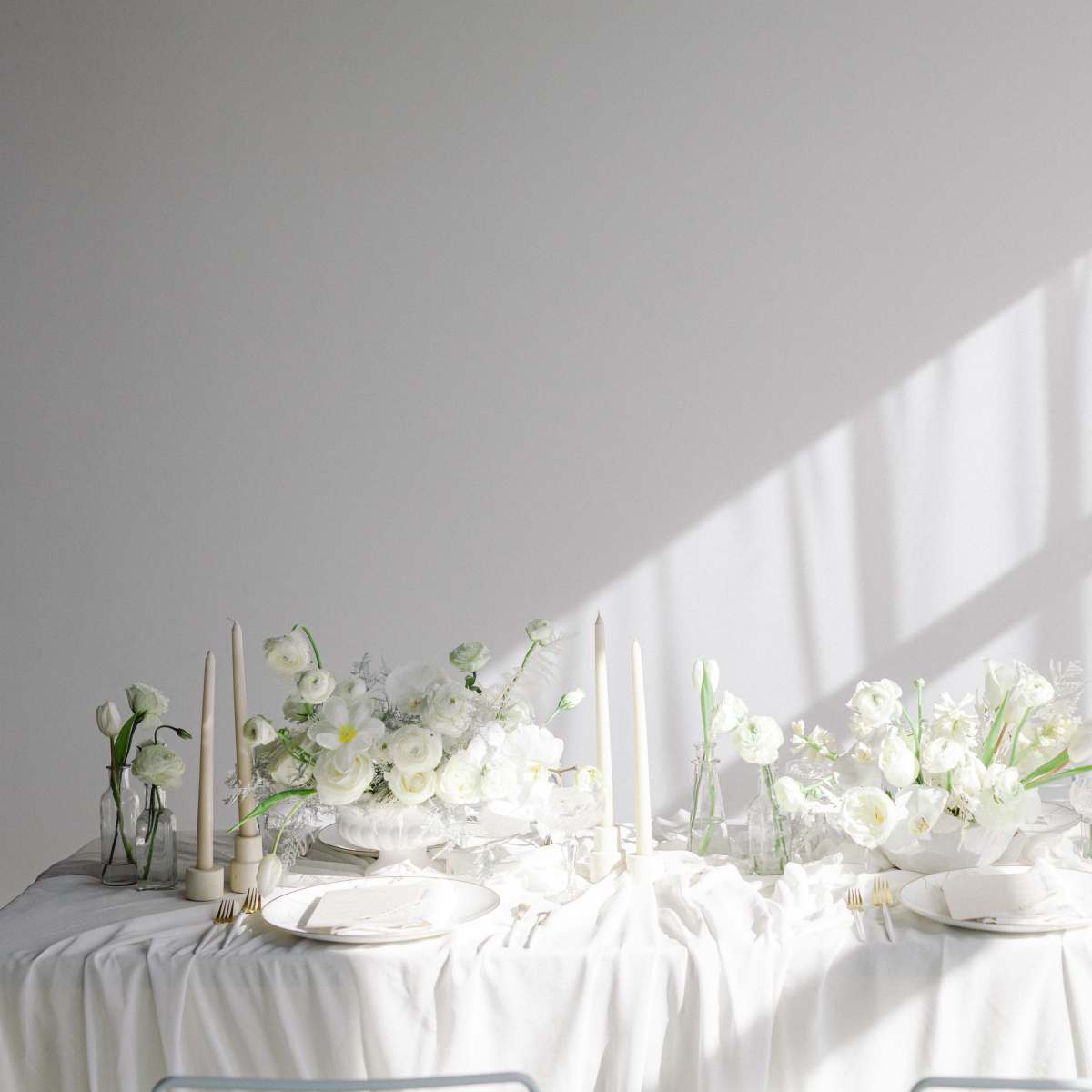A dining table is set with white tablecloths, floral centerpieces, candle holders, and plates under soft, natural light.