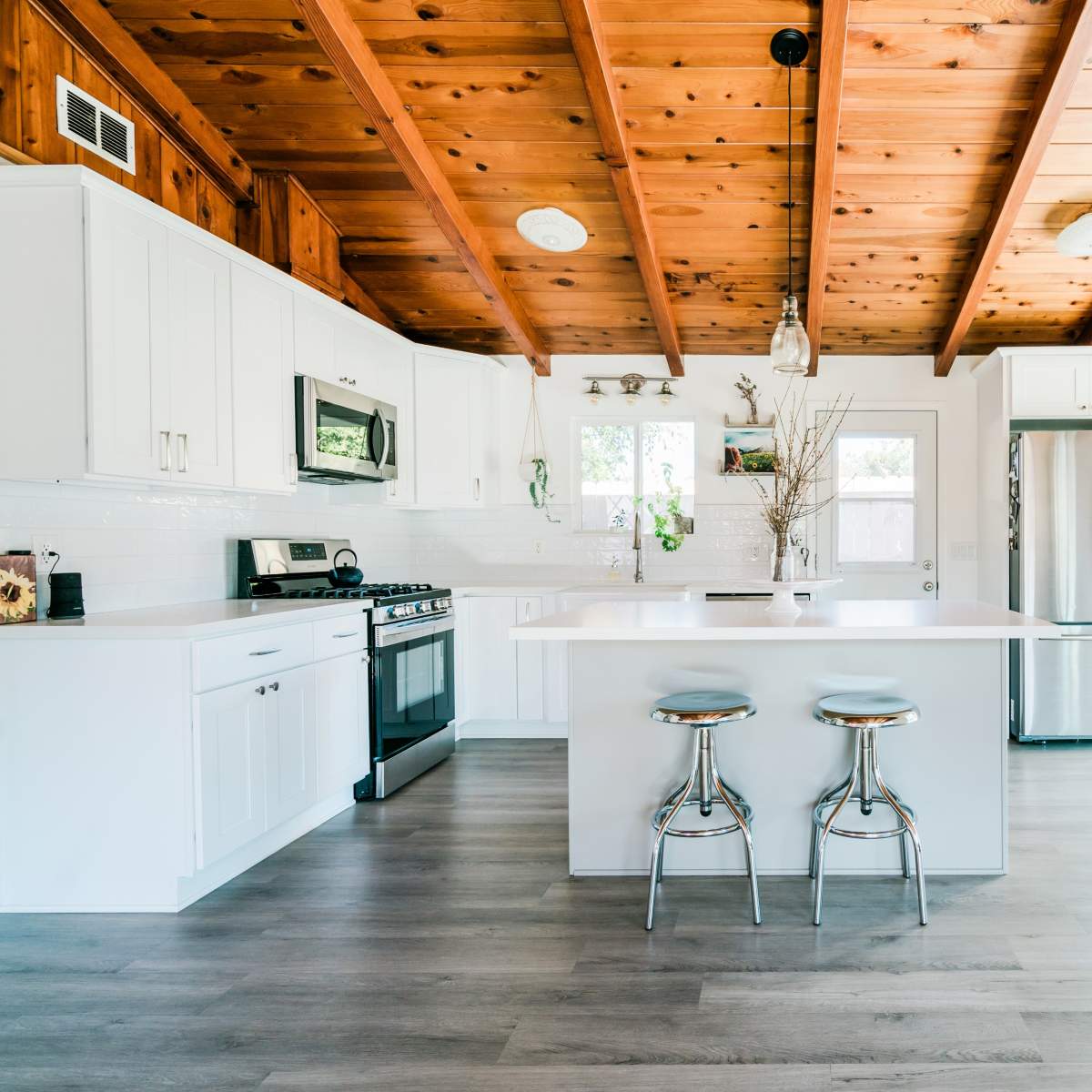 A modern kitchen with white cabinetry, a central island with two metal stools, and exposed wooden beams on the ceiling.