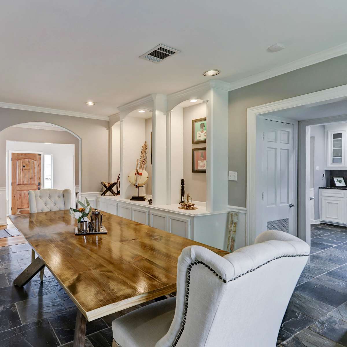 A wooden dining table with decorative items is centered in a well-lit room, framed by white wainscoting and gray walls, with doors leading to adjacent spaces.