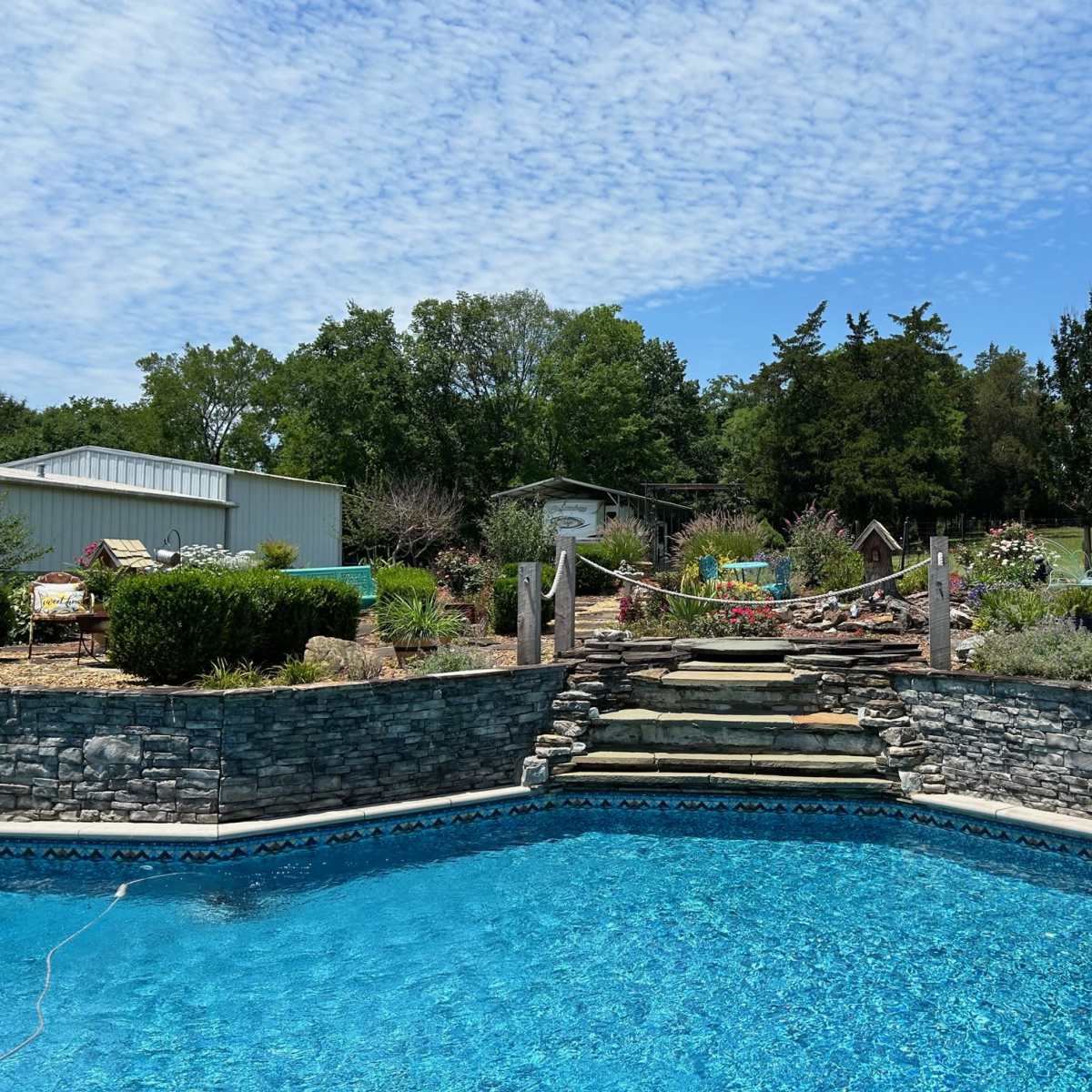 The scene shows a swimming pool surrounded by landscaped gardens and a building in the background under a cloudy blue sky.