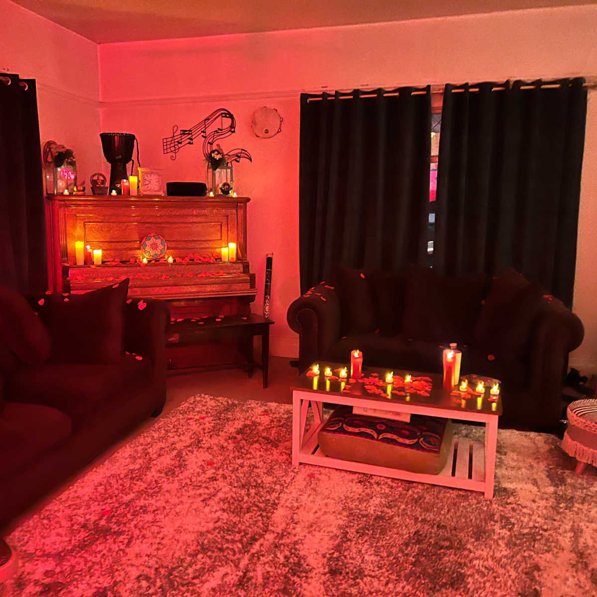 A dimly lit living room with dark curtains, a wooden piano adorned with candles, and two couches surrounding a coffee table.