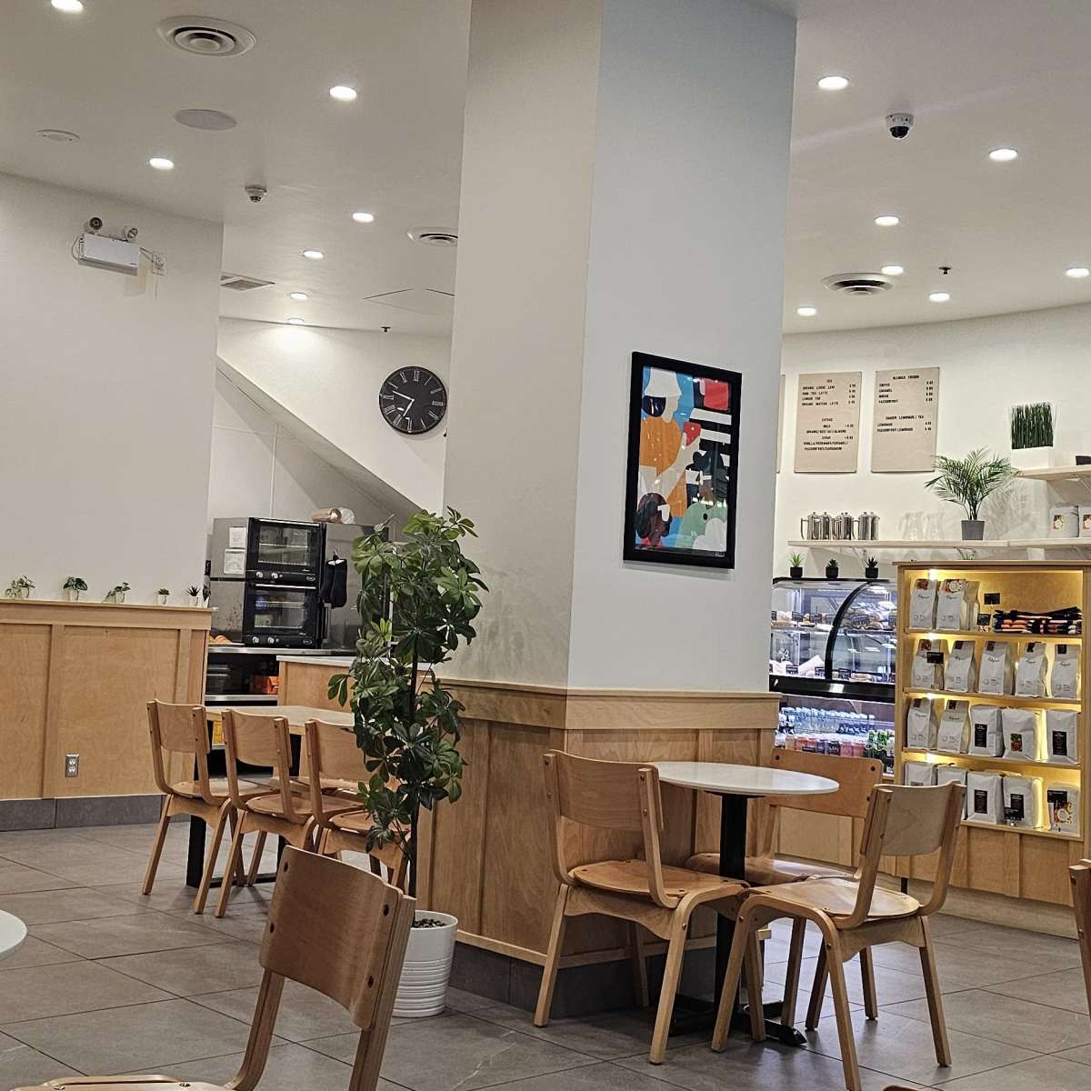 A modern café interior with light wood furniture, a coffee preparation area, and shelves displaying products.