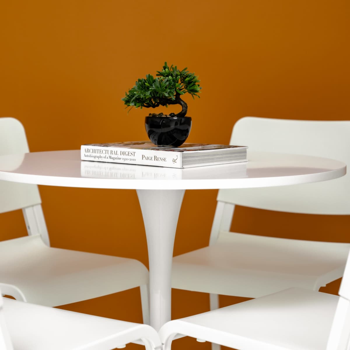 A round white table is surrounded by four white chairs, with a small potted plant and a stack of books on top.