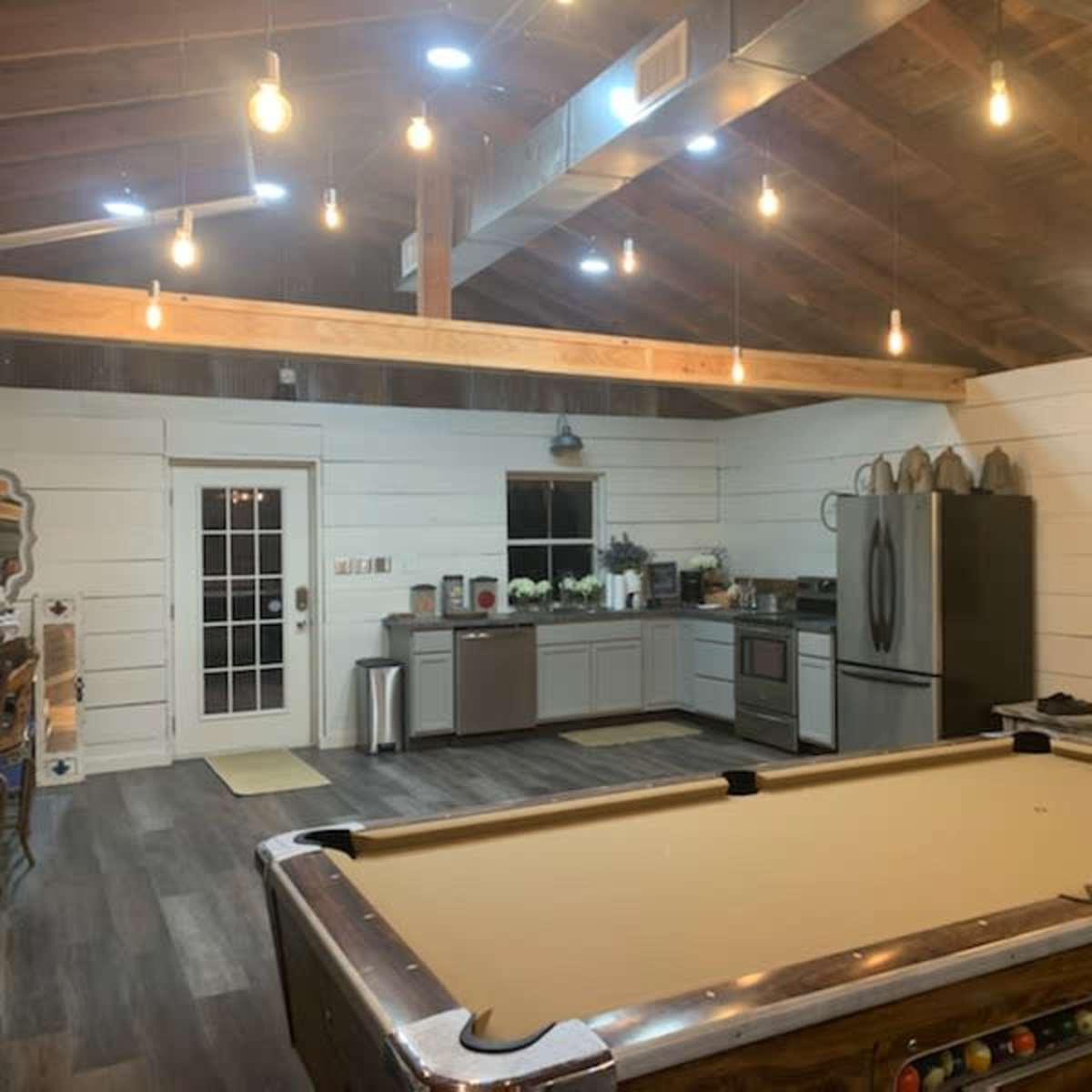 A spacious, well-lit room featuring a pool table, a modern kitchen with stainless steel appliances, and white shiplap walls.