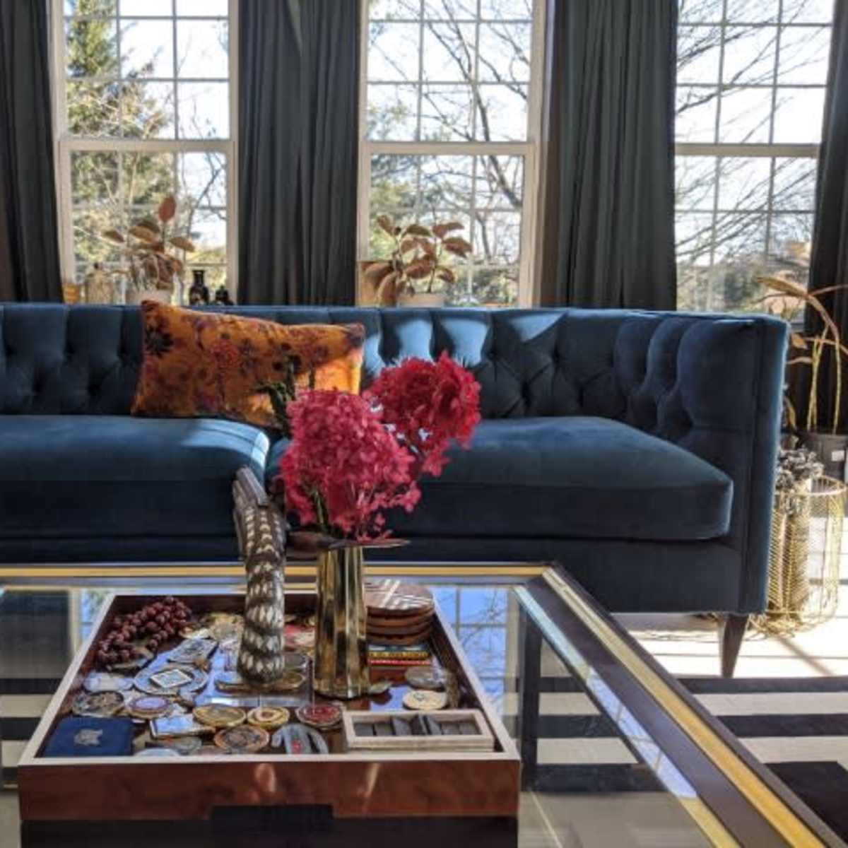 A blue tufted sofa with a decorative pillow, positioned in front of a coffee table adorned with flowers and various items, all set against large windows.